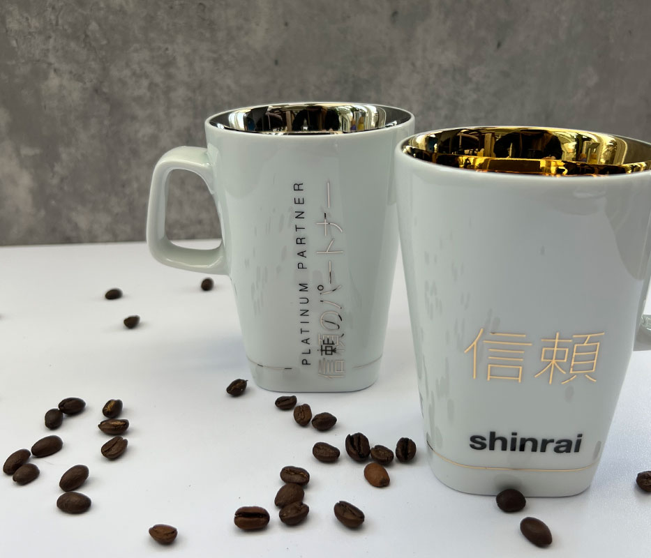 Zwei weiße Henkelbecher mit gold- und silberfarbenen Details zwischen Kaffeebohnen